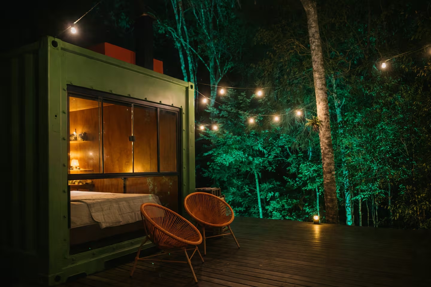 Cabana acesa à noite, com a cama vista pela janela e o deck iluminado