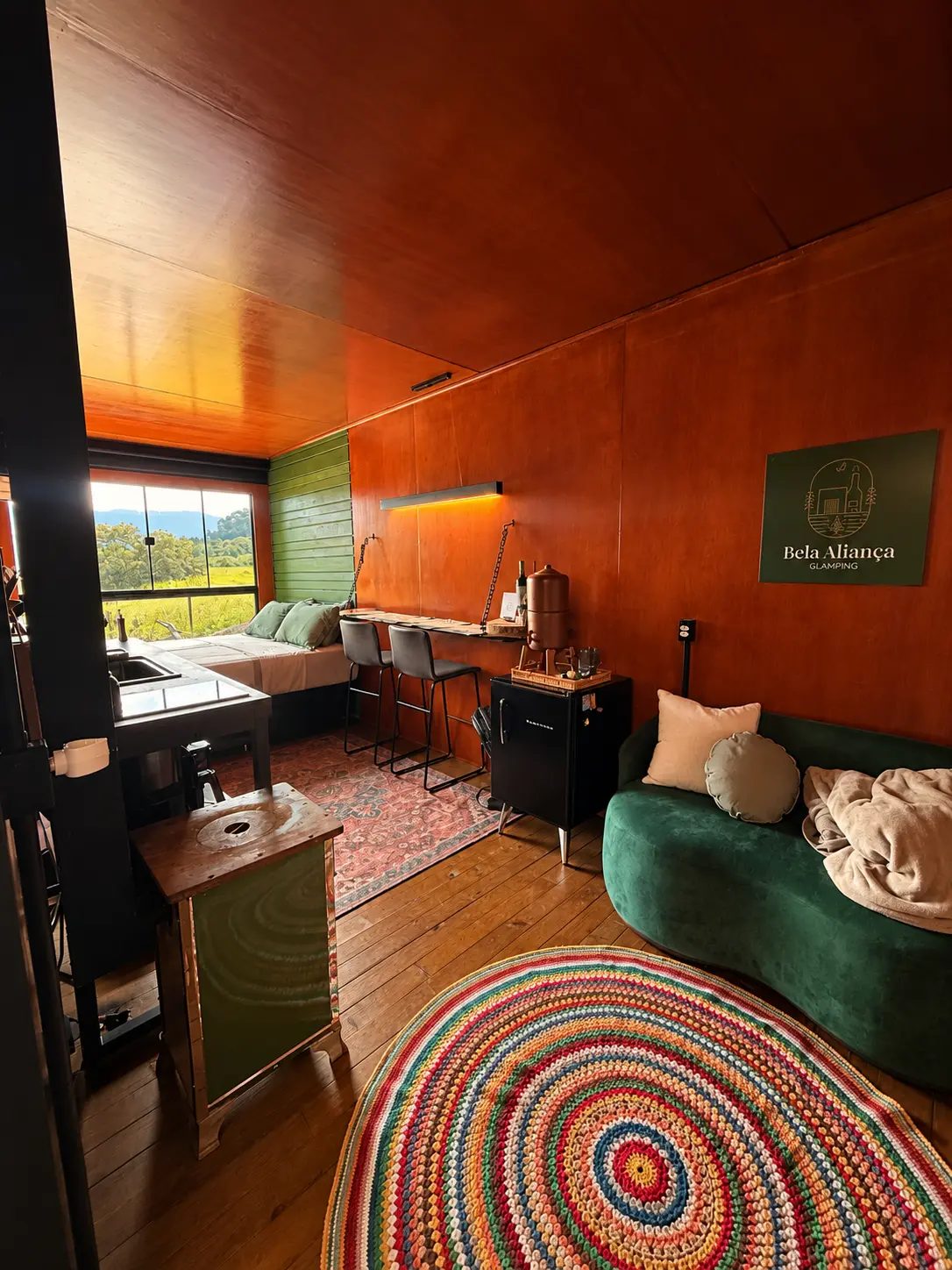 Sala de estar da cabana de dia, com sofá verde, rug colorida e a placa do Bela Aliança